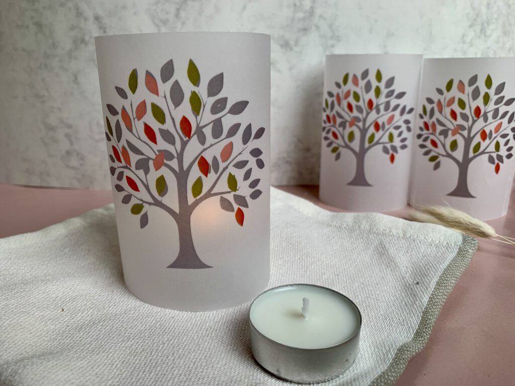windlicht-taufe-deko-kerze Detailaufnahme eines selbstgebastelten Windlichts als Tischdeko für die Taufe mit Lebensbaum-Motiv, Teelicht im Inneren, Teelicht daneben. Deko Ideen zur Taufe.