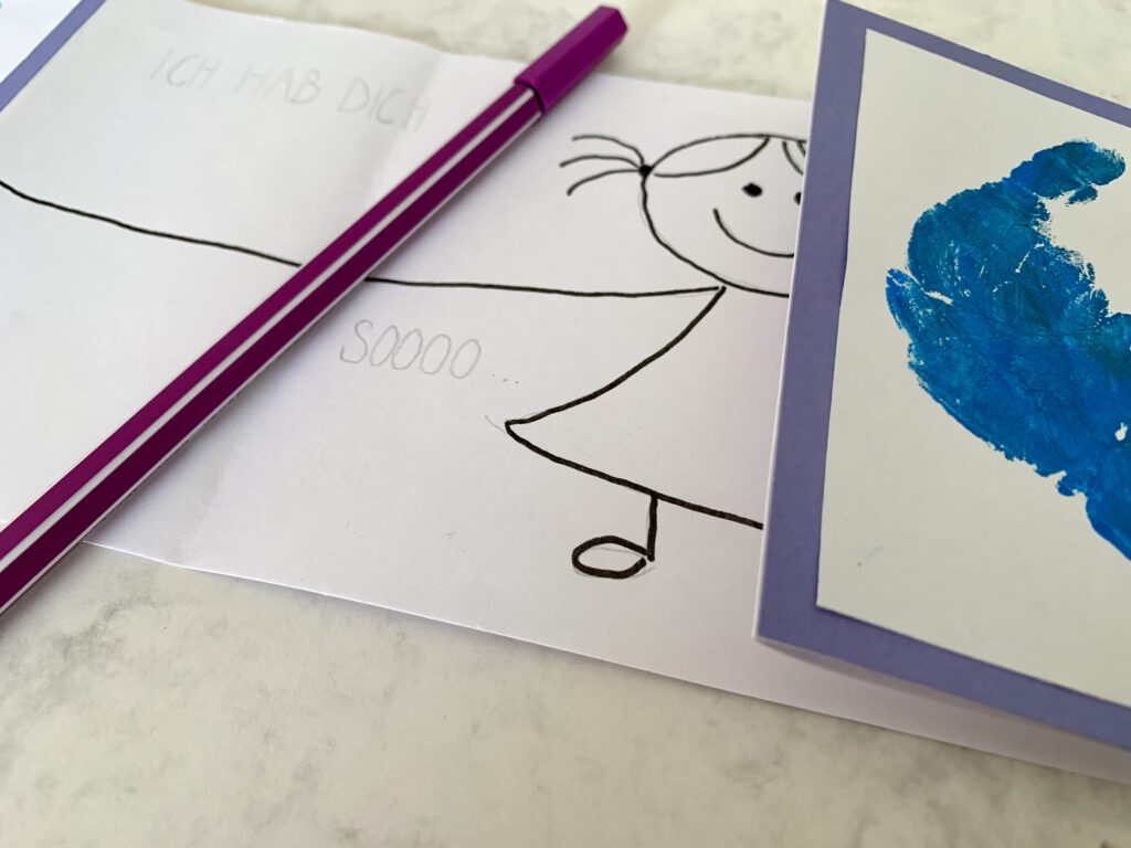 Karte_zum_Vatertag_basteln_mit_Handabdruck Nahaufnahme vom Bastelprozess. Wir basteln eine Karte zum Vatertag mit Handabdruck. Die Handabdrücke werden außen auf die Klappkarte geklebt. im Inneren wird eine süße Figur gezeichnet, die die Arme ausbreitet. Hier wird der Schriftzug "ich hab dich so liebt" mit Bleistift vorgezeichnet.
