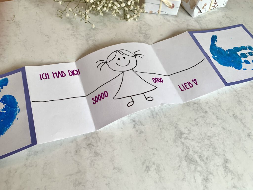 Vatertagskarte_basteln_mit_Baby Selbstgebastelte Vatertagskarte mit Handabdrücken von Kleinkind. Aufgeklappt und mit liebevoller Gestaltung im Inneren. Geschenke im Hintergrund.