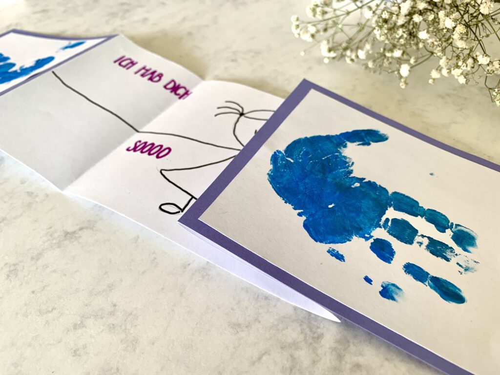 Vatertagskarte_mit_Handabdruck_Baby Halb geöffnete Vatertagskarte mit Einblick auf Handabdruck und Zeichnung im Inneren. Schriftzug ich hab dich so lieb. Bastelidee für Kleinkinder zum Vatertag