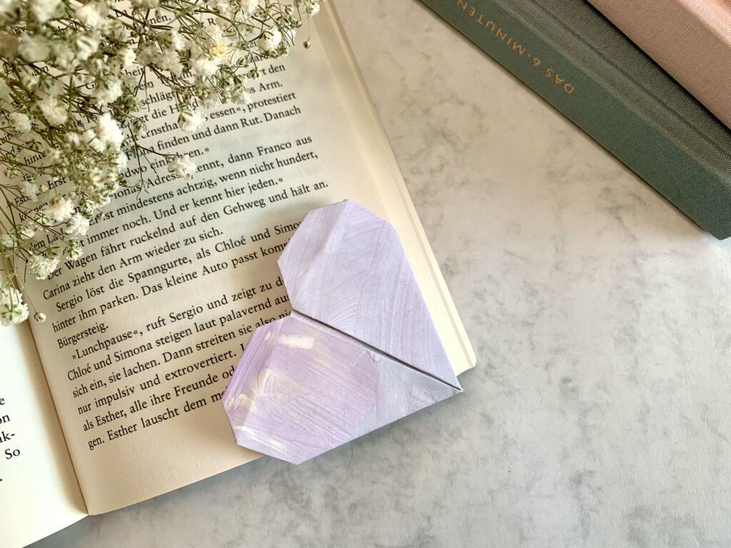basteln-mit-kind-lesezeichen Herzförmiges Lesezeichen aus Kinderzeichnung liegt in einem aufgeklappten Buch, im Hintergrund ein pastellfarbener Bücherstapel – schnelles Geschenk basteln mit Kleinkind