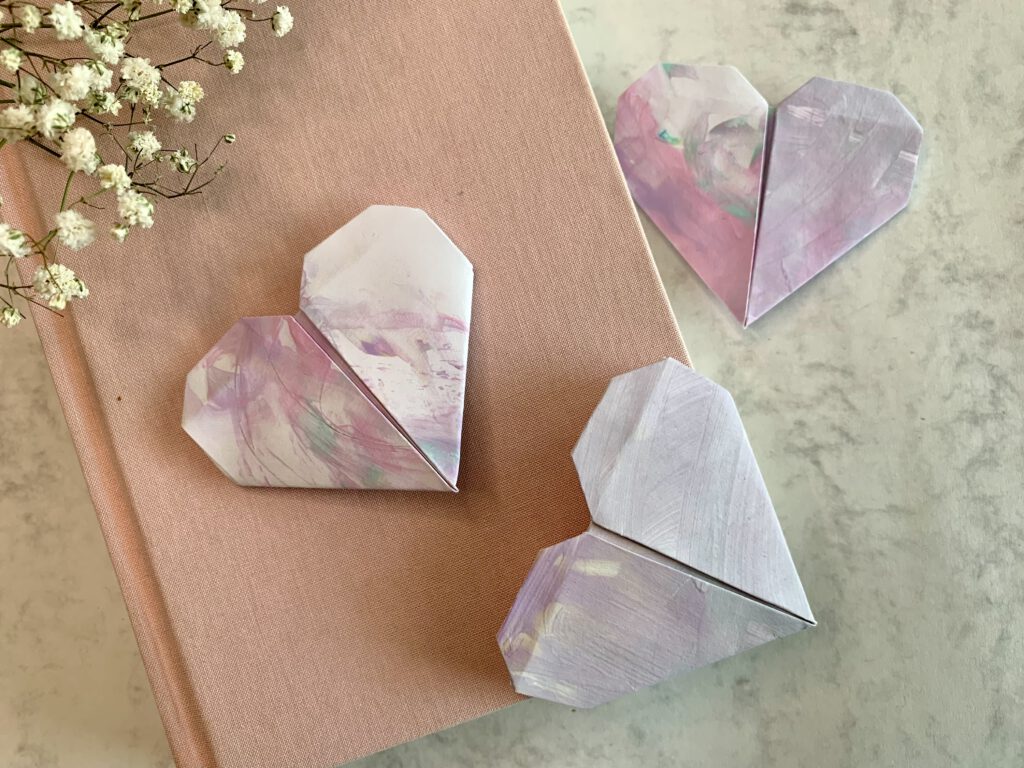 diy-geschenkidee-herz-lesen-kinderbild Geschenkidee mit Kindern: Lesezeichen nach Origamitechnik aus Kinderzeichnungen falten. 3 fertige Lesezeichen auf Buch mit rosa Einband, daneben Schleierkraut