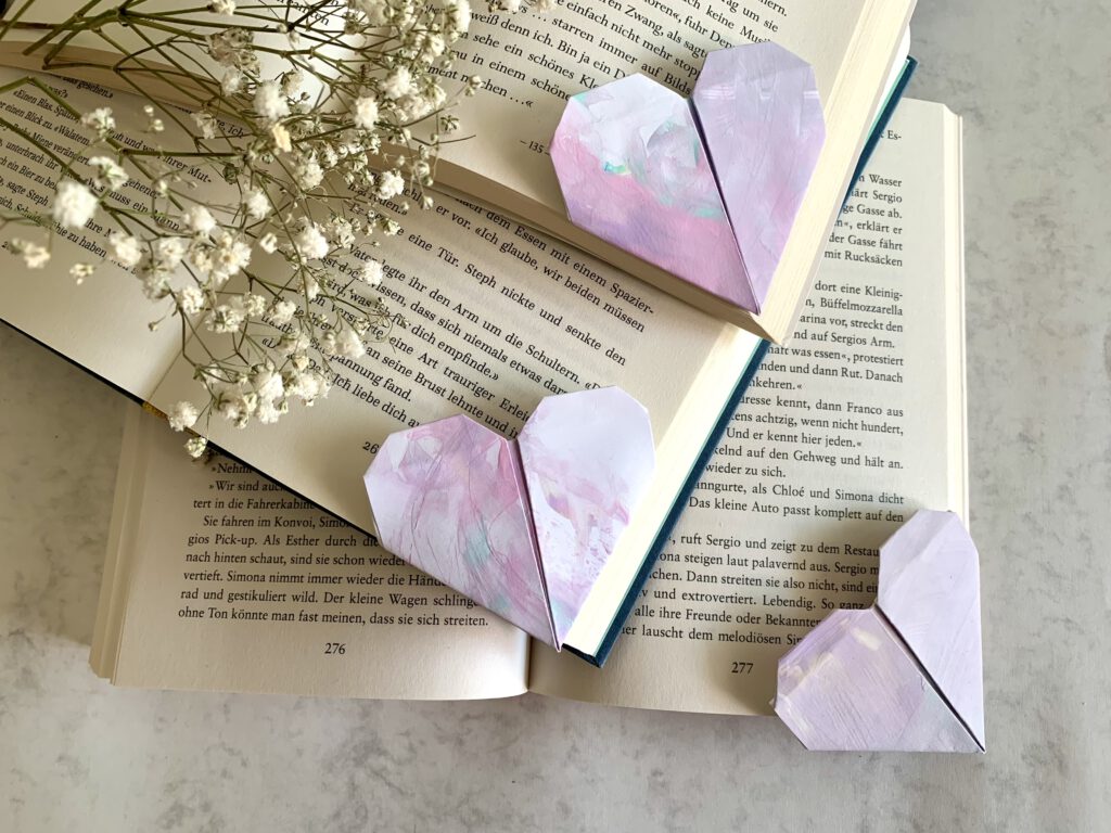Geschenkidee: Geschenk basteln mit Kindern. Aus Kinderzeichnungen falten wir herzförmige Lesezeichen. 3 fertige Lesezeichen auf aufgeschlagenen Büchern, Schleierkraut im Hintergrund
