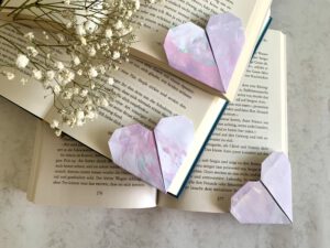 Geschenkidee: Geschenk basteln mit Kindern. Aus Kinderzeichnungen falten wir herzförmige Lesezeichen. 3 fertige Lesezeichen auf aufgeschlagenen Büchern, Schleierkraut im Hintergrund