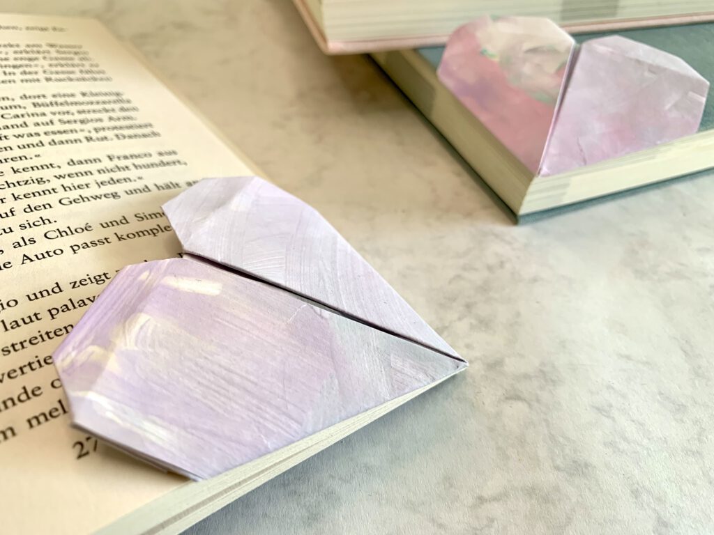 personalisiertes-lesen-herz-geschenk-kinderzeichnung Zwei selbst gebastelte Origami-Lesezeichen aus Kinderzeichnungen auf einem geöffneten Buch – kreative Bastelidee für Kinder ab 2 Jahren