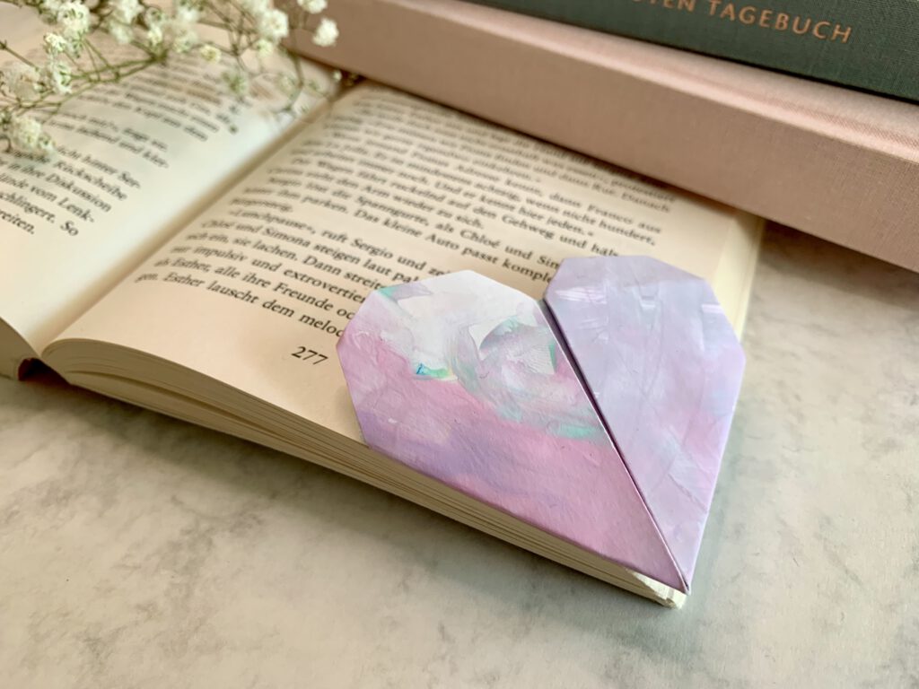sinnvolle-geschenke-mit-kindern-basteln Herzförmiges Lesezeichen aus Kinderzeichnung liegt in einem aufgeklappten Buch, im Hintergrund ein pastellfarbener Bücherstapel – liebevolle DIY Geschenkidee mit Kleinkind