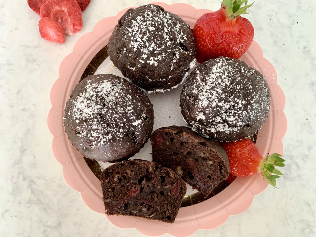 zuckerfreie-schokomuffins-mit-erdbeeren-aufgeschnitten Zuckerfreier Muffin auf einem Teller, in der Mitte aufgeschnitten – mit saftiger Schoko-Bananen-Textur, daneben frische Erdbeeren und weitere zuckerfreie Schokomuffins