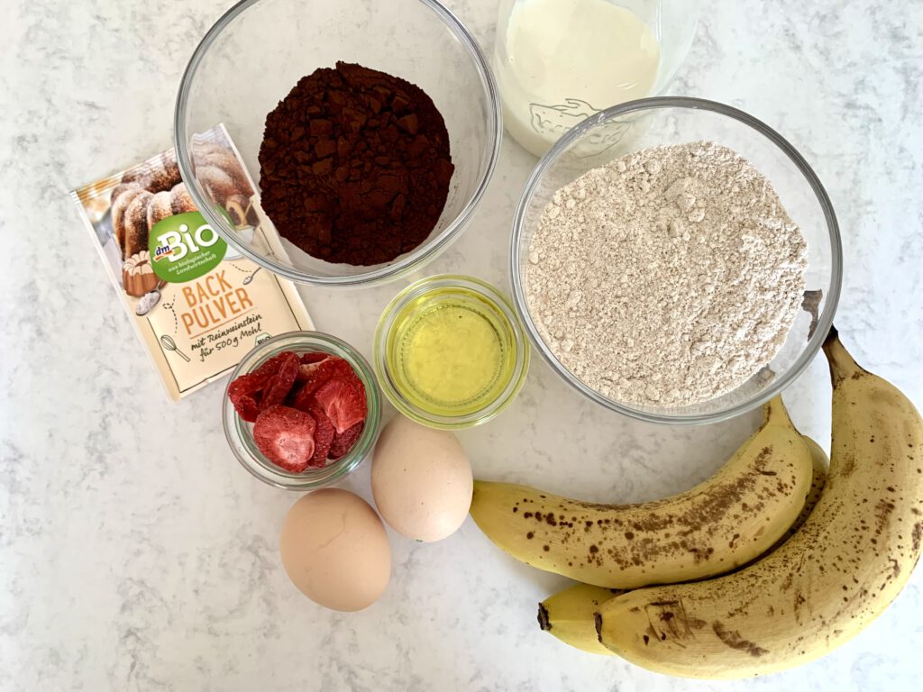 zuckerfreie-schokomuffins-zutaten Zutaten für zuckerfreie Schokomuffins, von oben fotografiert – mit Bananen, Mehl, Kakao, Eiern, Öl, Milch und getrockneten Erdbeeren auf einem Tisch