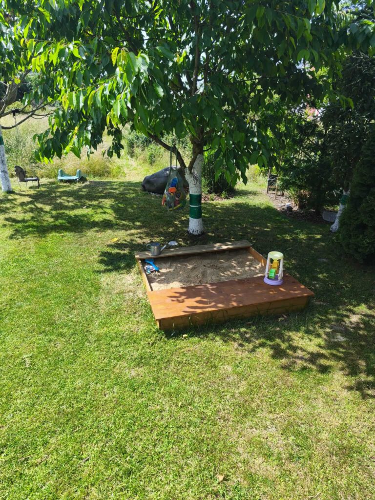 welchen-sandkasten-kaufen.jpg Kleiner Sandkasten im Garten. Sandkasten ist aus Holz mit Sitzmöglichkeiten.
