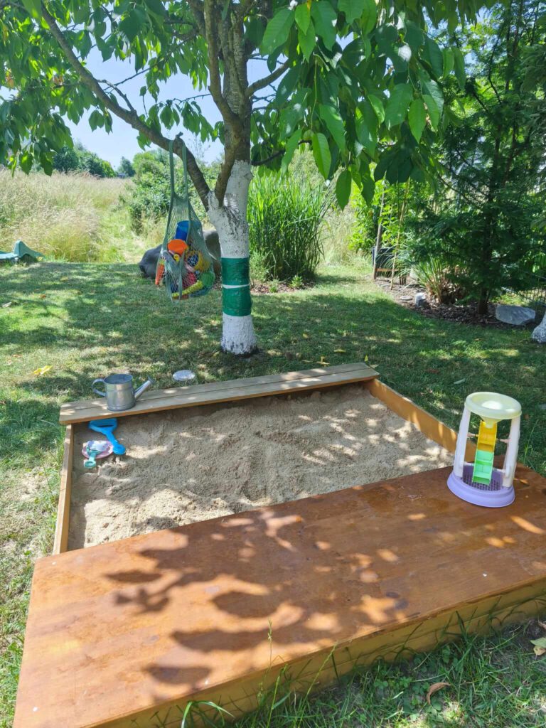 sandkasten-kaufen-empfehlung.jpg Sandkasten kaufen: Ein einfacher Holzsandkasten für Kleinkinder im Garten. Breite Sitzbänke, unter denen das Sandspielzeug verstaut werden kann.