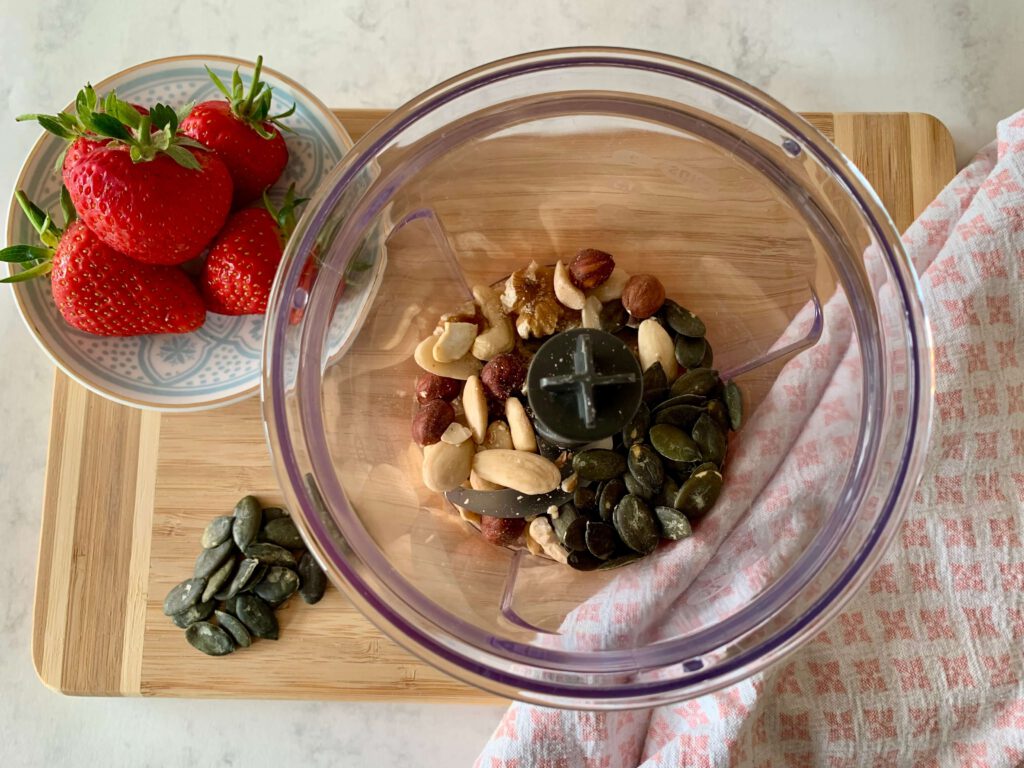 nuesse-kuerbiskerne-mixer-gesundes-fruehstueck-erdbeeren.jpg Zerkleinerte Nüsse und Kürbiskerne im Mixer als Basis für ein selbstgemachtes, proteinreiches Instant-Porridge. Im Hintergrund frische Erdbeeren auf einem Holzbrett – gesundes Frühstück vorbereiten.