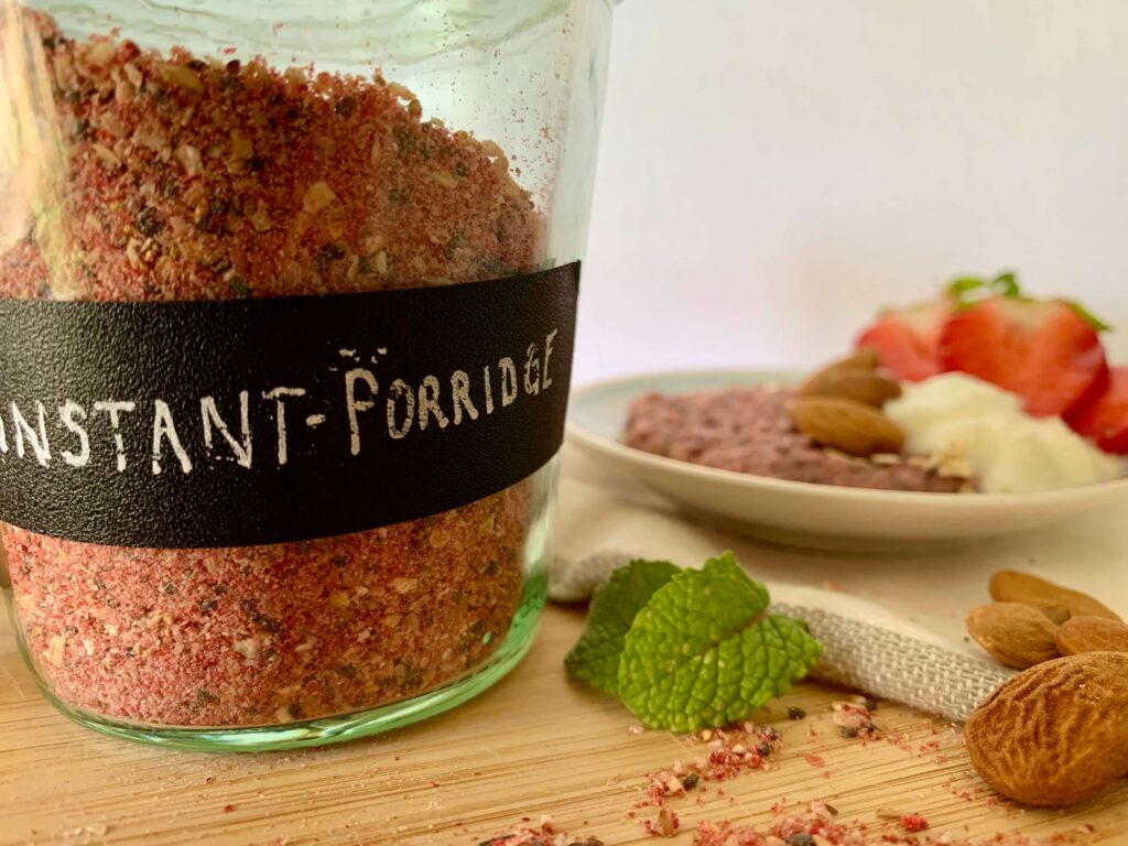instant-porridge-beschriftetes-glas-mit-topping-fruehstuecksidee.jpg Beschriftetes Schraubglas mit selbst gemachtem Instant-Porridge, im Hintergrund ein angerichtetes Frühstück mit frischen Toppings – ideal zum Vorbereiten.