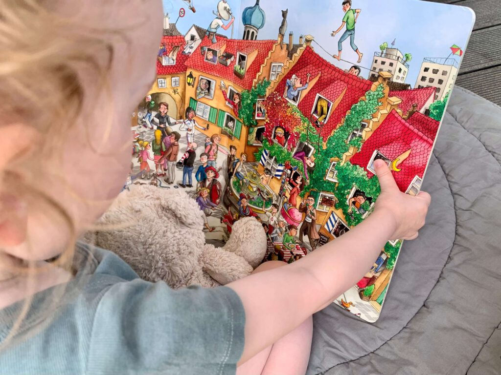 Kleinkind-wimmelbuch-augsburg.jpg Kleinkind schaut sich mit seinem Teddy das Bilderbuch "Das große Augsburger Wimmelbuch" an.