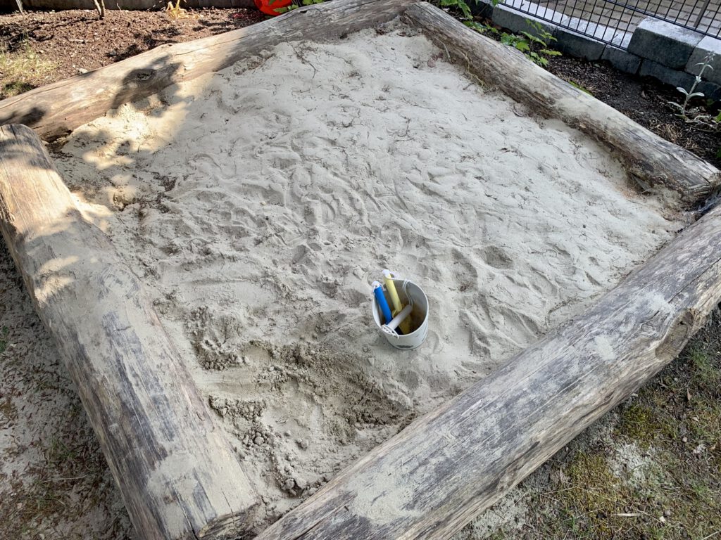 sandkasten-holz-baumstamm-einfassung-von-oben.jpg Großer Holzsandkasten mit massiver Einfassung aus Baumstämmen – Draufsicht auf das natürliche Modell im Garten.