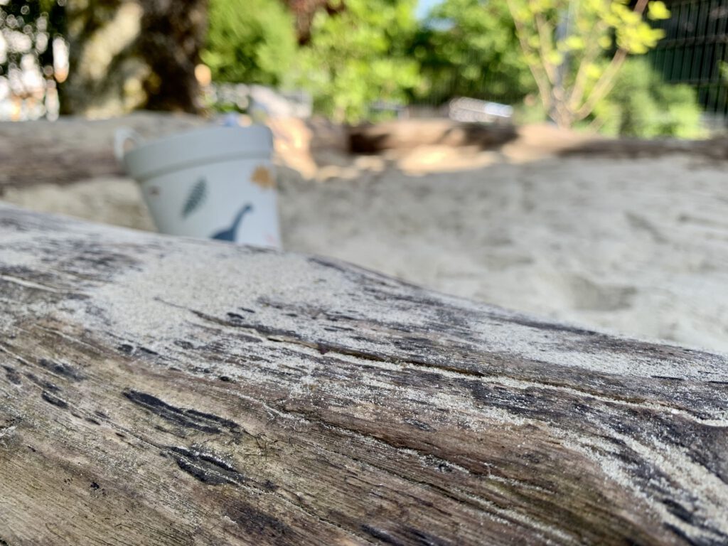 holzsandkasten-detail-balken-sandeleimer-garten.jpg Detailaufnahme eines robusten Holzsandkastens mit breiten Holzbalken; im Hintergrund steht ein bunter Sandeimer im Sand.