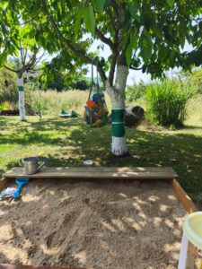 Welcher Sandkasten ist der beste? Holzsandkasten mit Sitzbank, im Hintergrund Sandspielzeug aufbewahrt in einem Einkaufsnetz