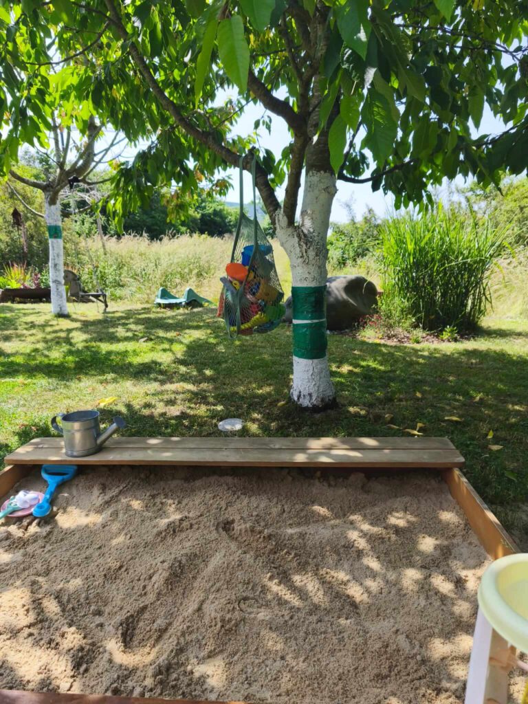 sandkasten-holz-mit-aufbewahrung.jpg Welcher Sandkasten ist der beste? Holzsandkasten mit Sitzbank, im Hintergrund Sandspielzeug aufbewahrt in einem Einkaufsnetz