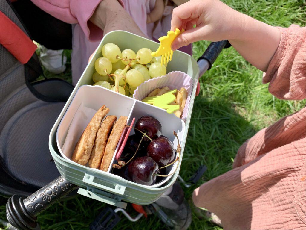 nackzeit im Freien: Kleinkind greift mit Pieksi & Griffel Pieksern zur Brotdose – Spaß beim Essen unterwegs.