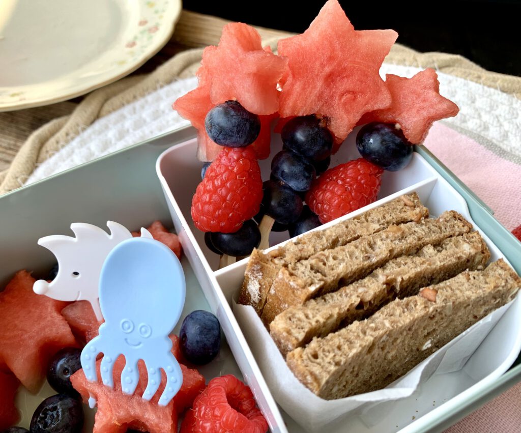 Ideen für die Kita-Brotdose: Vollkornbrot mit Aufstrich, Zauberstäbe aus Obst und Snack Piekser von Pieksi und Griffel. Die Obstspieße bestehen aus Melone, Blaubeeren und Himbeeren.