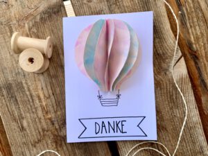 Eine selbstgebastelte Dankeskarte für die Kindergarten Erzieherin: Ein 3D-Heißluftballon aus Kinderzeichnungen in rosa und hellblau, darunter das Wort DANKE