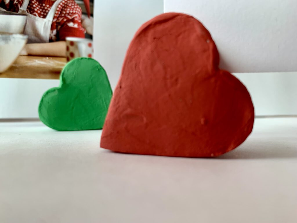 Herzförmiger roter Fotohalter aus Modelliermasse, Detailaufnahme mit grünem Fotohalter im Hintergrund. Ideal als Geschenk und um Erinnerungen in Szene zu setzen.