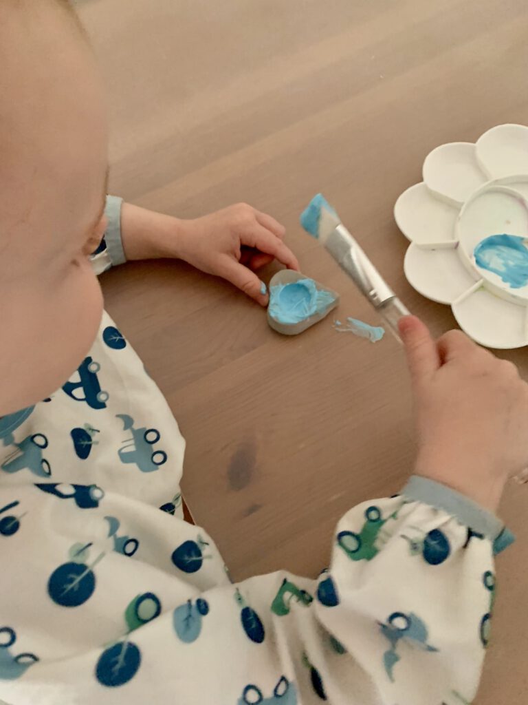 Zweijähriges Kind bemalt herzförmigen Kerzenhalter aus Modelliermasse mit blauer Fingerfarbe – kreative Bastelidee für Kleinkinder.