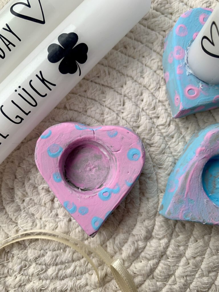 Kerzenhalter in Herzform, bemalt mit Kinderfingerfarben, dekoriert mit weißen Kerzen und Schriftzügen als DIY Geschenkidee.
