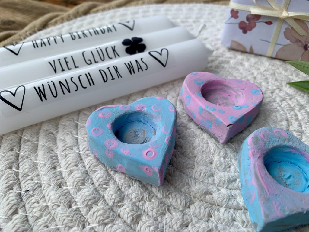 Mehrere herzförmige Kerzenhalter, bemalt in Rosa und Hellblau, mit bedruckten Kerzen und Kerzentattoos. Aufschriften: ‚Wünsche dir was‘, ‚Viel Glück‘, ‚Happy Birthday‘