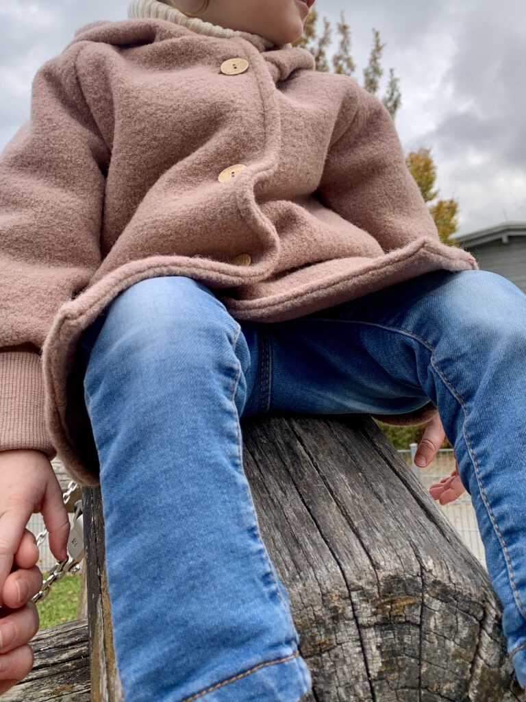 Kleinkind sitzt auf dem Klettergerüst und trägt die Wollwalkjacke von Petit Cochon in Puder – warm und bequem.