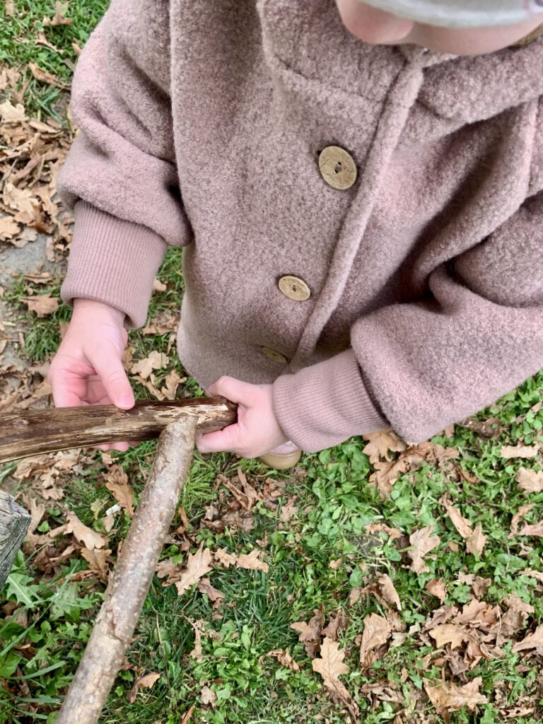 Kleinkind spielt mit Stöcken im Herbstlaub und trägt die Wollwalkjacke von Petit Cochon – robuste, natürliche Kinderkleidung.