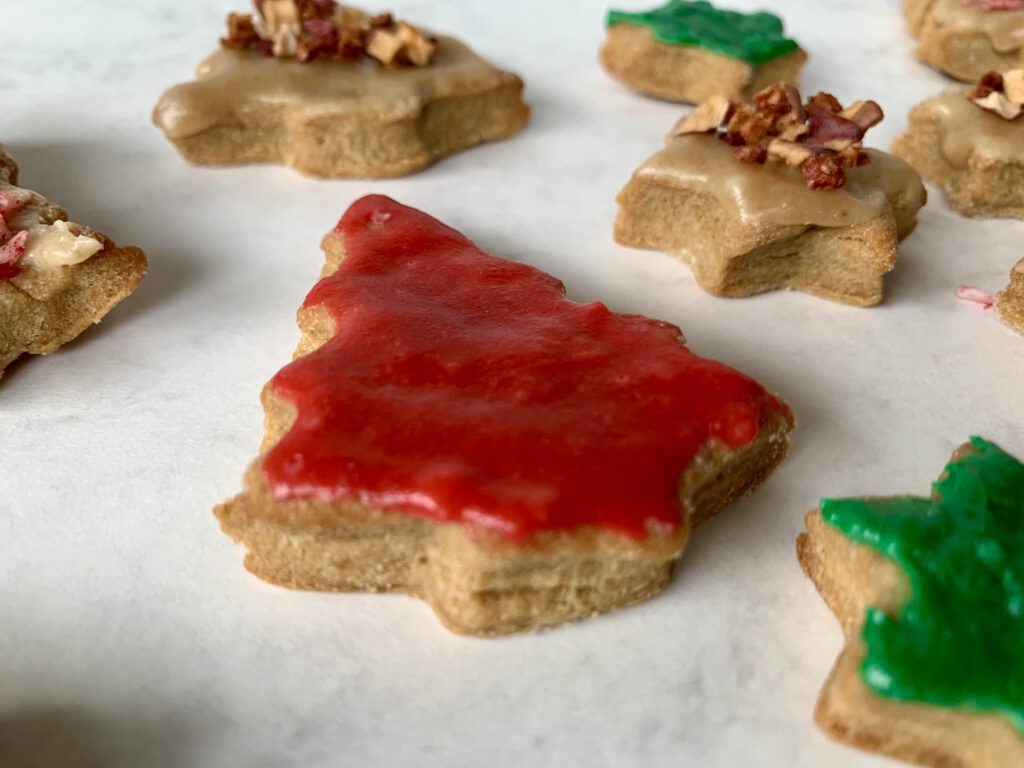 Tannenbaumplätzchen mit rotem Mandelmus-Zuckerguss, im Hintergrund Sterne mit buntem Fruchtstreuseldekor