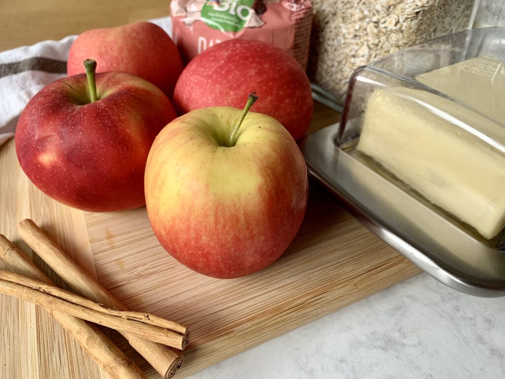 Zutaten für einen zuckerfreien Apfel Crumble mit Äpfeln, Haferflocken, Mehl, Butter und Dattelsüße auf einer Arbeitsfläche.