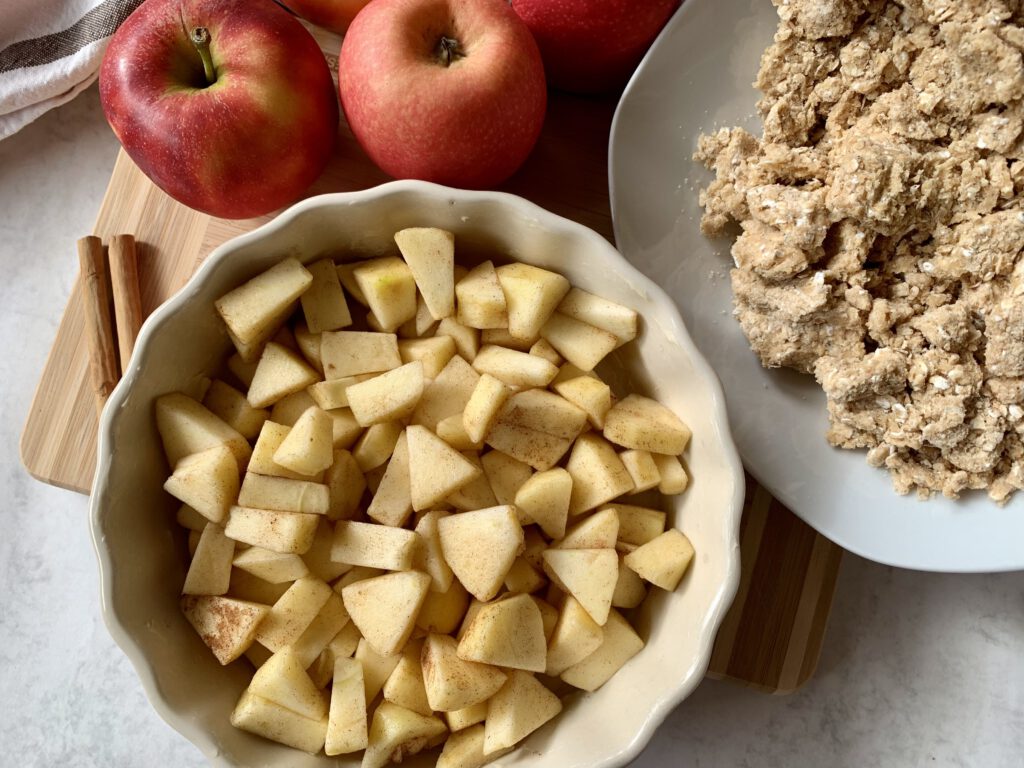 Zubereitung eines Apple Crumbles ohne Zucker: Apfelstücke mit Zimt und Dattelsirup sowie Schüssel mit Streuselteig.