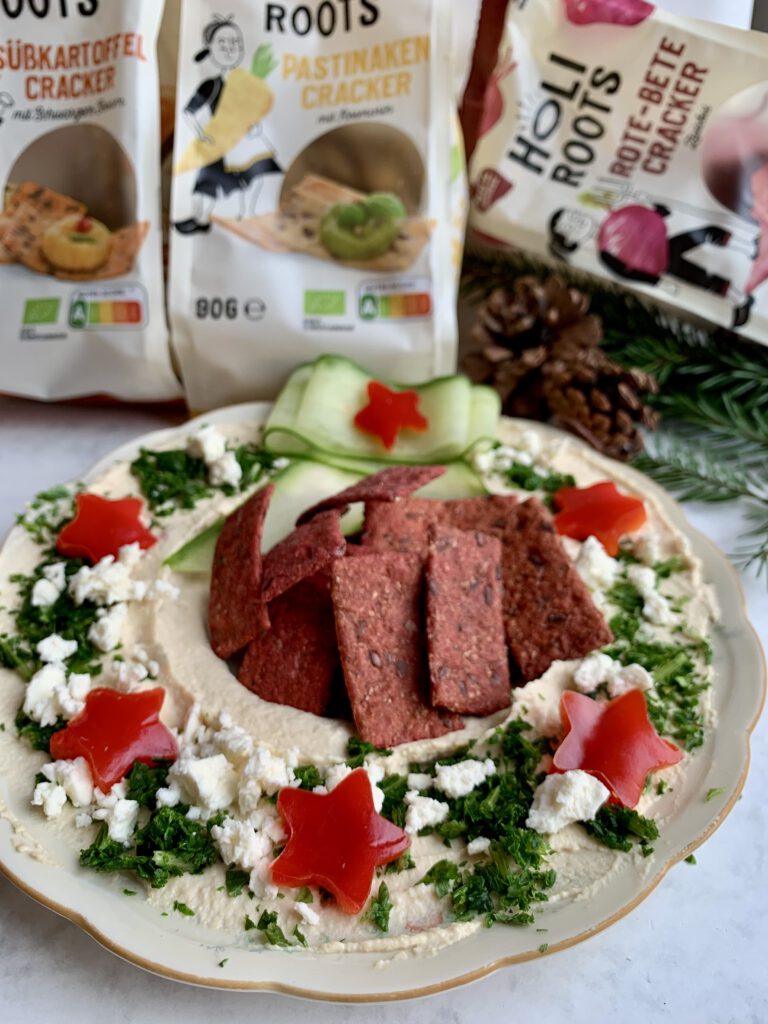 weihnachtliche-snackplatte-holiroots-cracker.jpg Weihnachtliche Snackplatte mit Holiroots Crackern, dekoriert für die Feiertage, mit Crackerpackungen im Hintergrund. Hummus-Kranz schnell gemacht.