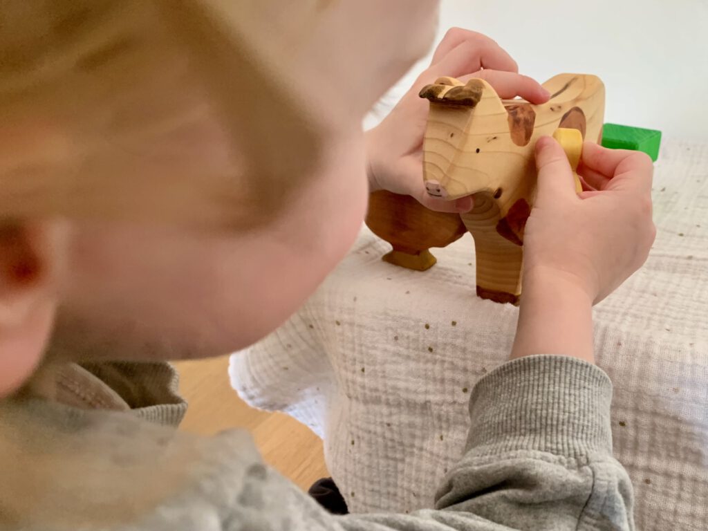 Kleinkind spielt mit einer Sägefisch-Holzkuh und einem Küken – ideales Holzspielzeug für offenes Spiel ab 3 Jahren.