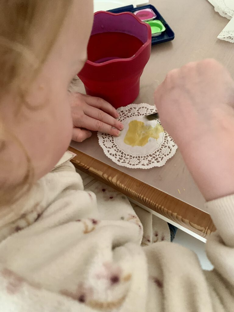 kind-bemalt-tortenspitze-wasserfarben-weihnachtsbasteln.jpg Alt-Text: Kleinkind bemalt eine Mini-Tortenspitze mit Wasserfarben für eine einfache Weihnachtsbastelidee.