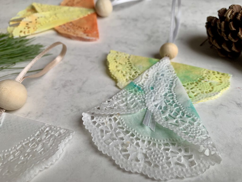 engel-basteln-nahaufnahme-tortenspitze-band-holzperle.jpg Nahaufnahme eines selbstgebastelten Weihnachtsengels aus Tortenspitze. Die Tortenspitze wurde von Kleinkindern bemalt. In wenigen Minuten falten wir daraus bezaubernde Engel.