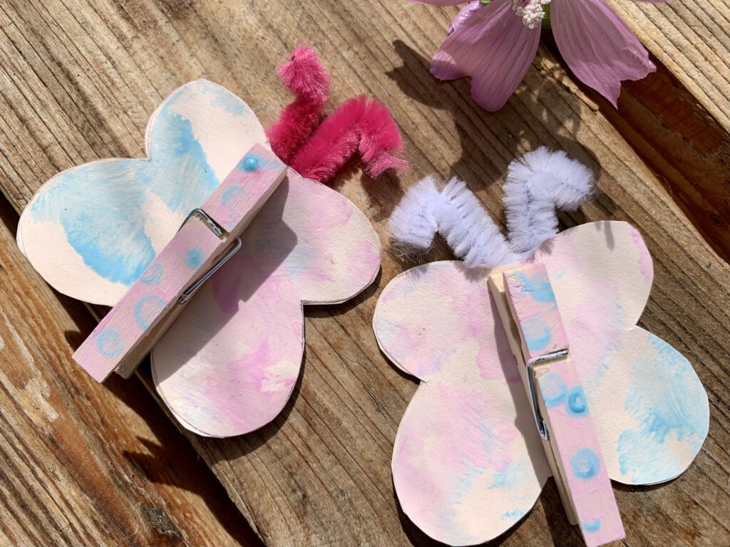Zwei Schmetterlinge in Pastellfarben. Wäscheklammern und Tonpapier werden mit Fingerfarben bemalt. Flügelform aus Tonpapier ausgeschnitten und zusammen mit Pfeifenputern als Flügel hinter die Wäscheklammer geklebt. Eine schnelle Bastelidee mit Kleinkind für den Frühling oder Sommer.