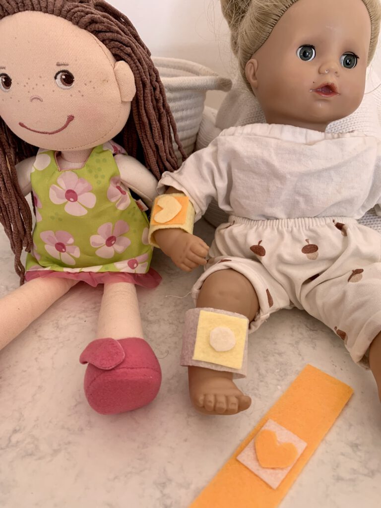 Zwei Puppen mit selbstgemachten Spielpflastern aus Filz mit Herz und Punkt, im Vordergrund liegt ein Puppenpflaster mit Herz