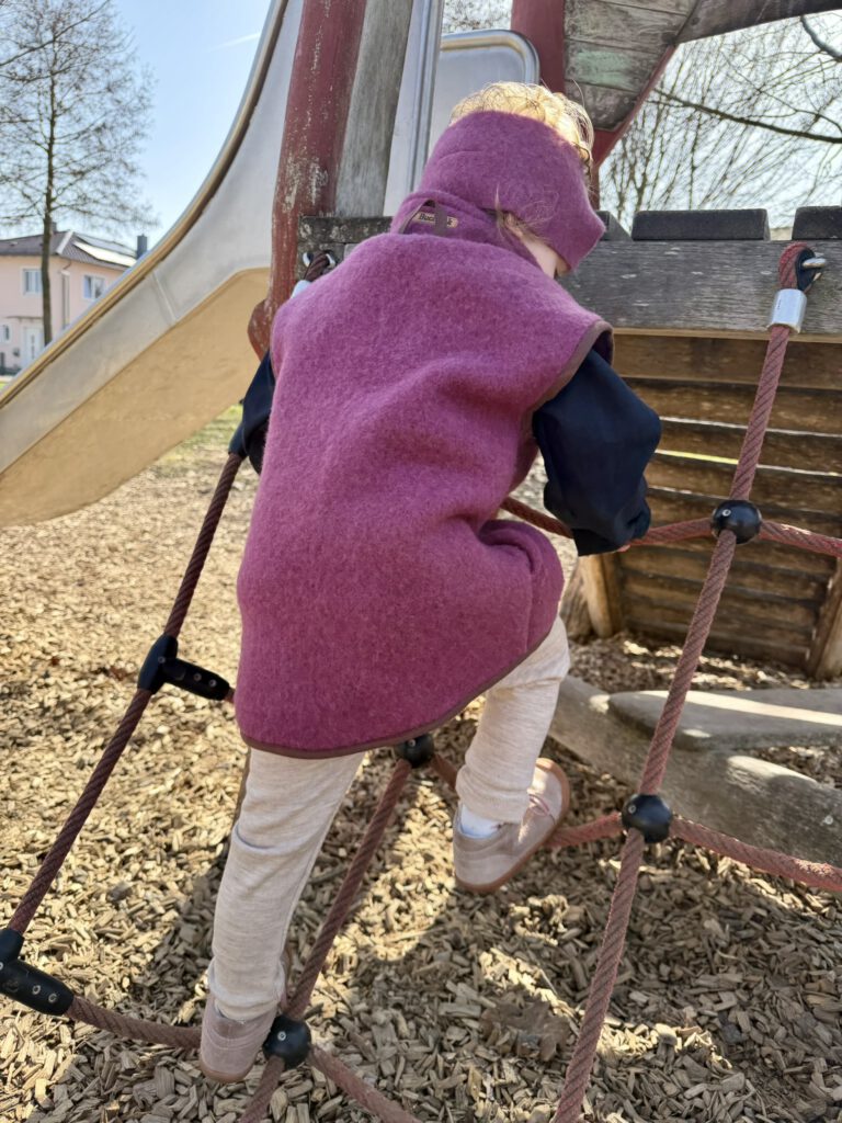 Kind beim Klettern auf dem Spielplatz mit Wollfleeceweste, Stirnband und nachhaltigem Frühlingsoutfit
