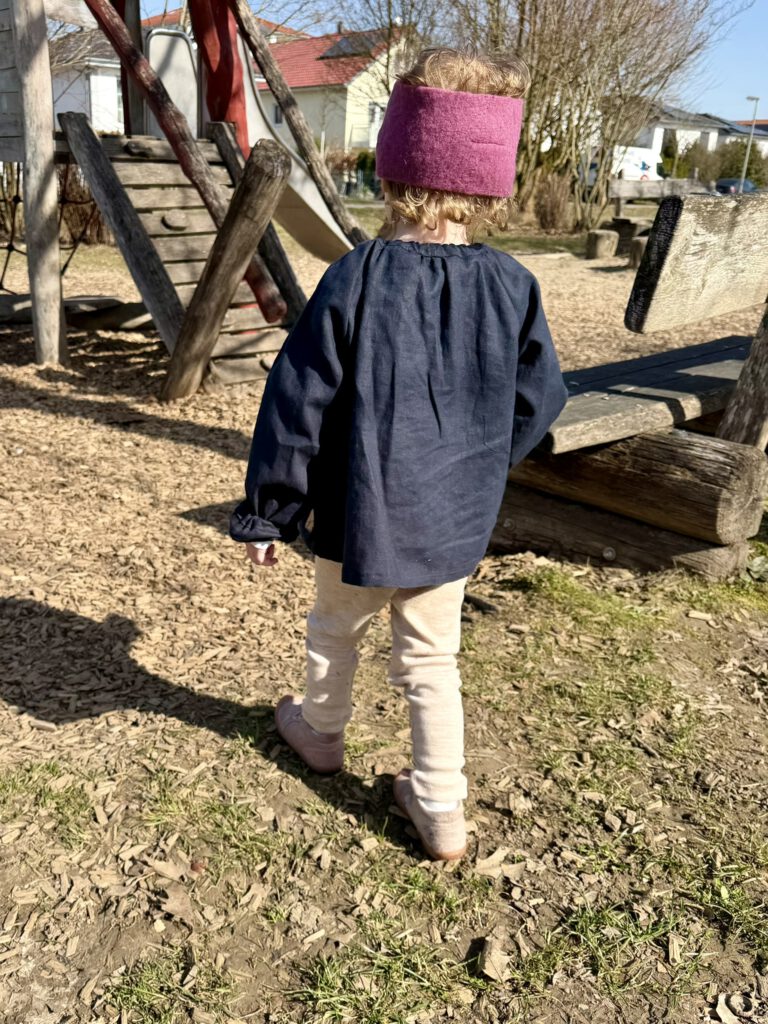 Kind von hinten im Frühling auf dem Spielplatz mit Stirnband, Leinenbluse und Wollleggings aus Naturmaterialien