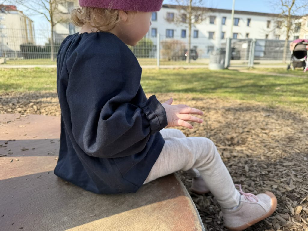 Kleinkind auf dem Spielplatz mit dunkelblauer Leinenbluse und Wollleggings